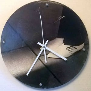 May include: Circular wall clock featuring a monochrome image of a shoe. The clock face has white hands and silver fixings. The shoe is light-coloured, set against a dark, textured background.