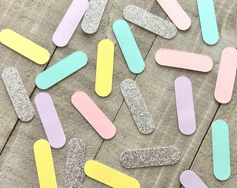 May include: A collection of pastel-colored confetti in the shapes of long, thin rectangles. The confetti is scattered on a light brown wooden surface. The colors include light pink, light blue, light purple, yellow, and silver.