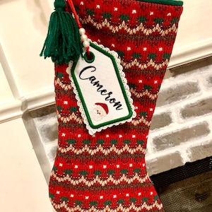 Puede incluir: Calcet&iacute;n de Navidad rojo y verde con una etiqueta blanca que dice "Cameron". El calcet&iacute;n tiene un borla verde y una cuenta blanca.