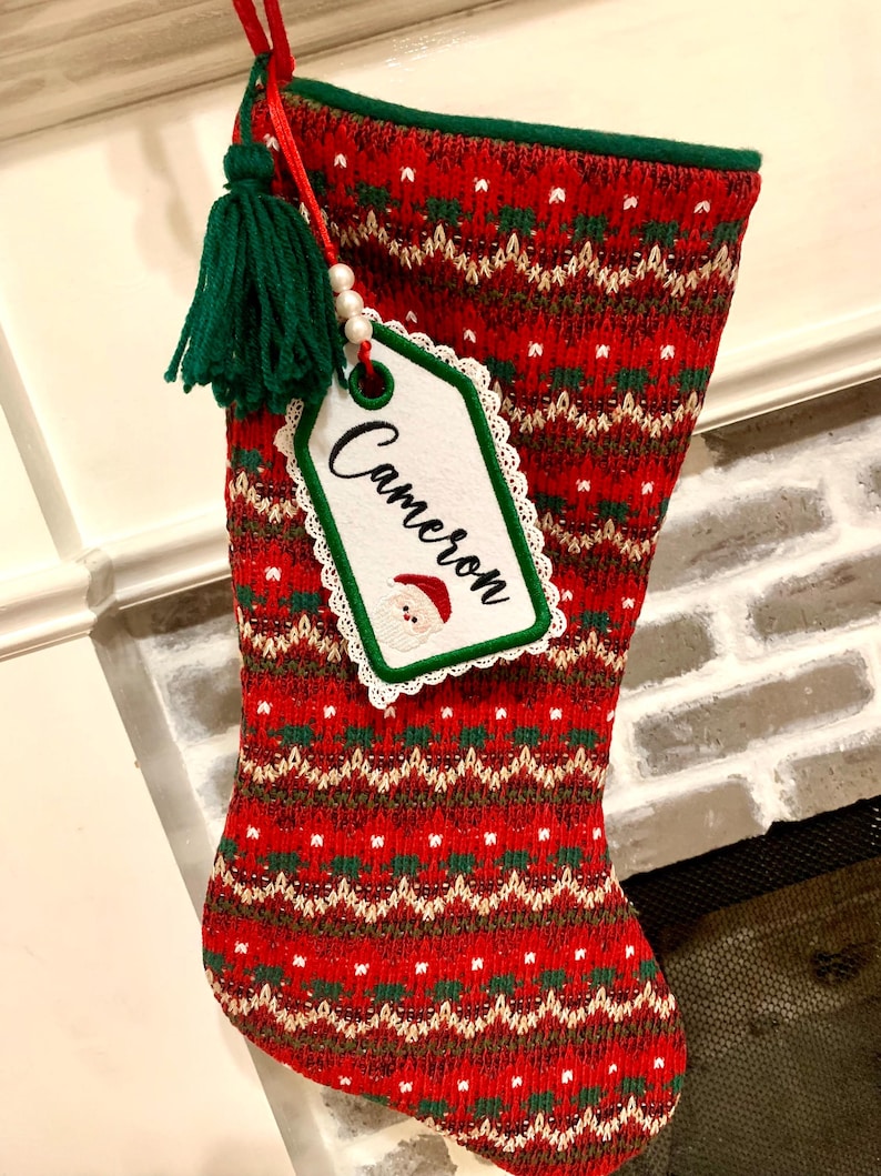 Puede incluir: Calcet&iacute;n de Navidad rojo y verde con una etiqueta blanca que dice "Cameron". El calcet&iacute;n tiene un borla verde y una cuenta blanca.