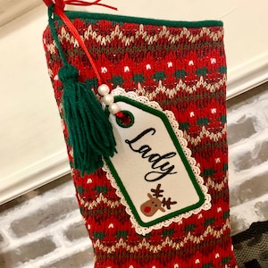 Puede incluir: Calcet&iacute;n de Navidad de punto rojo y verde con una etiqueta blanca que dice "Lady" y una ilustraci&oacute;n de un reno. El calcet&iacute;n tiene un borla verde.