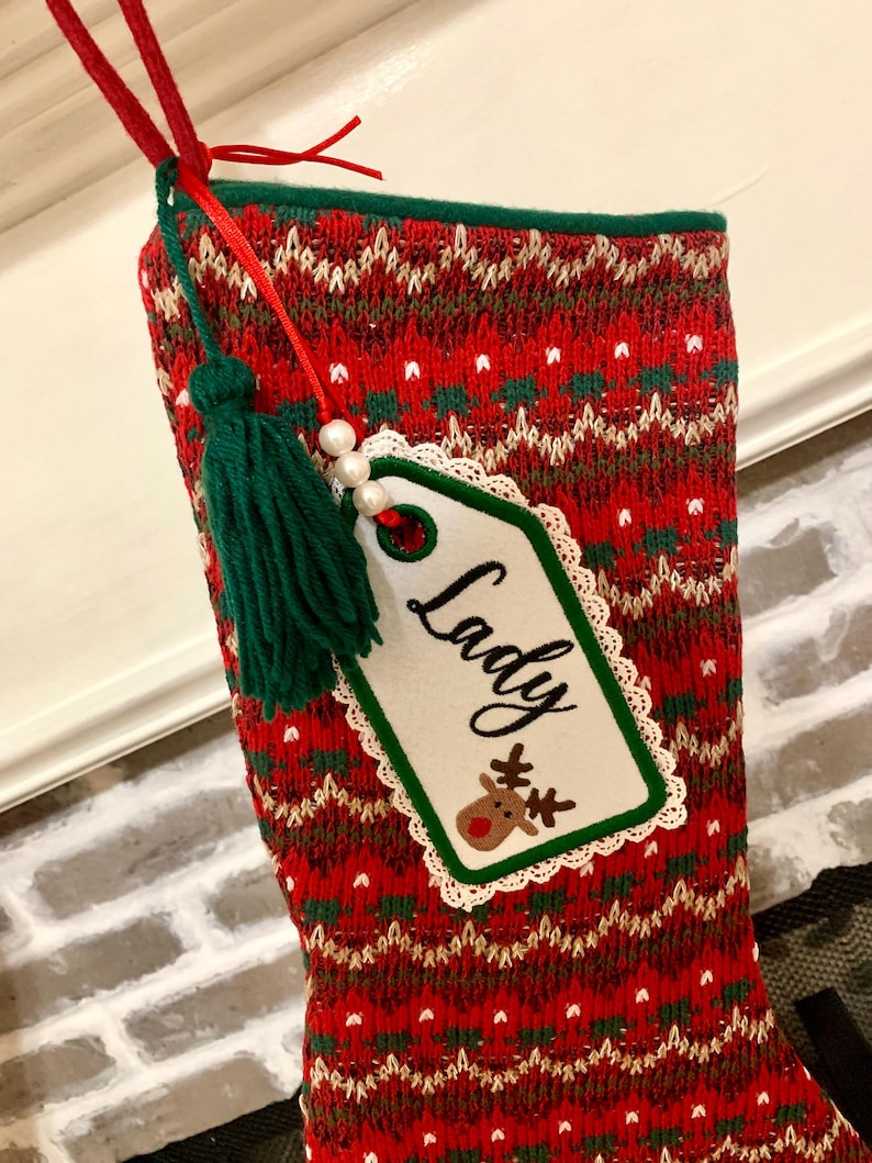 Puede incluir: Calcet&iacute;n de Navidad de punto rojo y verde con una etiqueta blanca que dice "Lady" y una ilustraci&oacute;n de un reno. El calcet&iacute;n tiene un borla verde.