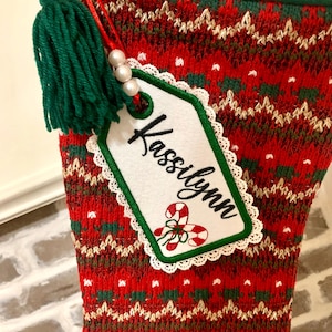 Puede incluir: Calcet&iacute;n de Navidad de punto rojo y verde con una etiqueta blanca que dice "Kassilynn" y un borla verde.