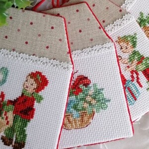 Può includere: Set di otto etichette regalo di Natale in tessuto bianco con bordo rosso e immagini ricamate di bambini e scene festive. Ogni etichetta ha un bordo di pizzo bianco e uno spago rosso.