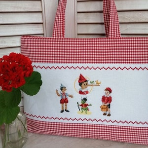 Può includere: Una borsa tote a quadretti rossi e bianchi con ricamo a punto croce. La borsa presenta un pannello bianco con personaggi ricamati di una storia. I manici e il bordo superiore sono a quadretti rossi e bianchi.