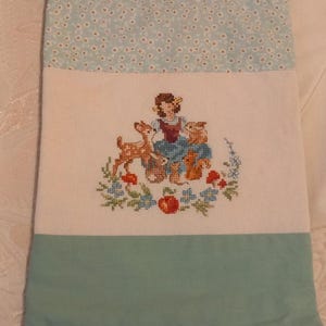 Può includere: Una borsa in tessuto blu chiaro e bianco con coulisse e fondo verde. La borsa presenta un ricamo a punto croce con una ragazza con due cervi e una mela rossa.