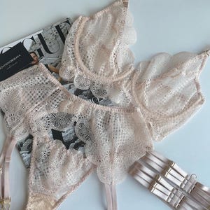 Puede incluir: Un delicado conjunto de lencería de encaje rosa claro, que incluye sujetador, bragas y liguero. El sujetador y las bragas presentan bordes festoneados. El conjunto se muestra sobre una revista con el texto "NOVODVORSKAYA".