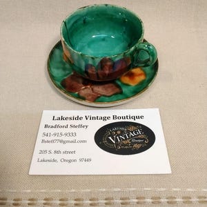 May include: A handcrafted ceramic teacup and saucer set. The cup and saucer are glazed in shades of green with brown and orange floral accents. The Lakeside Vintage Boutique business card is also in the image.