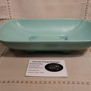 May include: A vintage, light turquoise soap dish with a rectangular shape and rounded corners. The dish has a raised center section and rests on four small feet. The Lakeside Vintage Boutique business card is in the foreground.