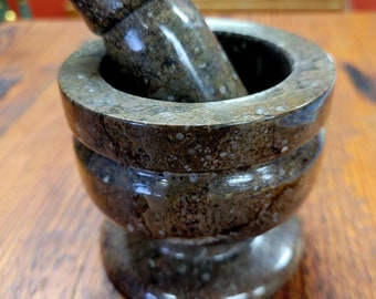 May include: A brown and gray stone mortar and pestle set. The mortar is round and has a wide base. The pestle is cylindrical and fits inside the mortar.