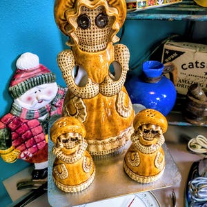 May include: A collection of vintage ceramic figurines, including a large doll-like figure and two smaller salt and pepper shakers. The figurines are in shades of tan and brown, with button eyes and a textured surface. A vintage scale is in the foreground.