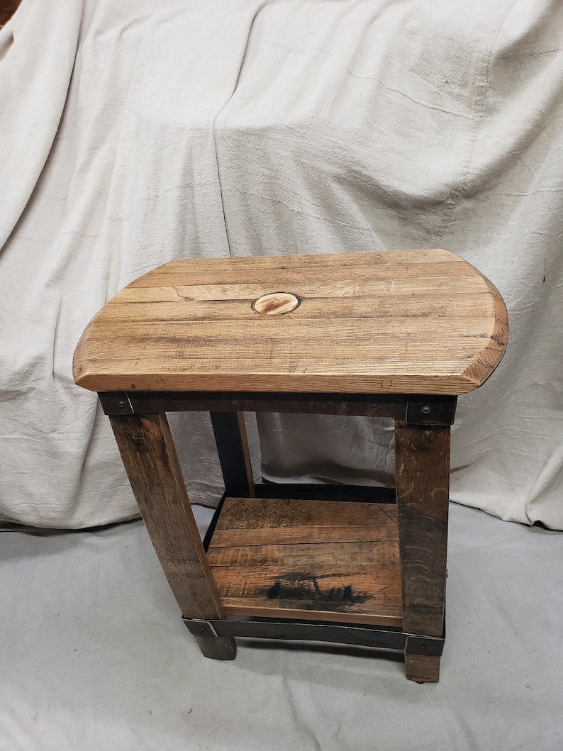 Pu&ograve; includere: Un tavolino rustico in legno con finitura naturale. Il tavolo presenta un piano arrotondato con un intarsio ovale e un ripiano inferiore. Le gambe angolate sono rinforzate con fasce metalliche, creando un aspetto industriale.