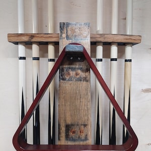 May include: A wooden wall-mounted rack for storing pool cues. The rack has a red wooden triangle rack for holding the cue ball and a shelf for storing other items. The rack is made from reclaimed wood and has a rustic look.