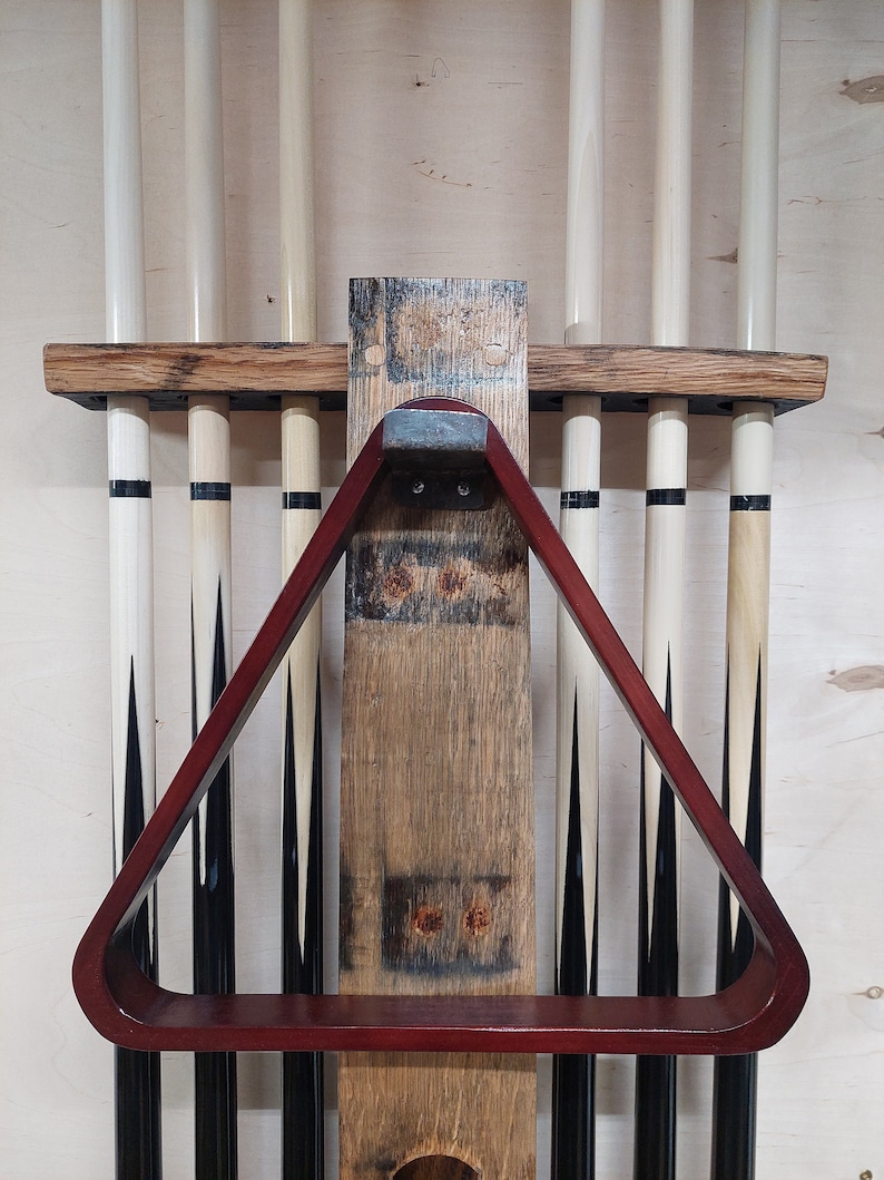 May include: A wooden wall-mounted rack for storing pool cues. The rack has a red wooden triangle rack for holding the cue ball and a shelf for storing other items. The rack is made from reclaimed wood and has a rustic look.