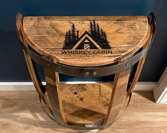 May include: A half-barrel table with a wooden top and metal frame. The top has a black and white logo that says "Whiskey Cabin" with a cabin and trees design.