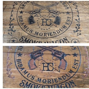 May include: Close-up of a wooden barrel with a black and white logo. The logo features two crossed revolvers, the letters "HC", and the text "YOWS BIBAMUS MORIENDUM EST SMOKE VAGON".