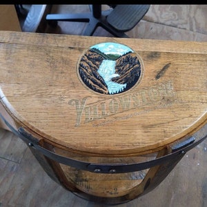 K&ouml;nnte beinhalten: Ein halbkreisf&ouml;rmiger Holztisch aus einem Fass mit einem dunklen Metallband am Rand. Die Oberfl&auml;che zeigt eine kreisf&ouml;rmige Grafik eines Wasserfalls in einer felsigen Landschaft mit dem Text Yellowstone Kentucky Straight Bourbon Whiskey.