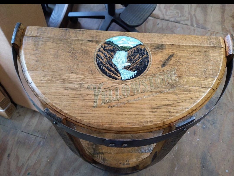 K&ouml;nnte beinhalten: Ein halbkreisf&ouml;rmiger Holztisch aus einem Fass mit einem dunklen Metallband am Rand. Die Oberfl&auml;che zeigt eine kreisf&ouml;rmige Grafik eines Wasserfalls in einer felsigen Landschaft mit dem Text Yellowstone Kentucky Straight Bourbon Whiskey.