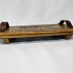 May include: A rustic wooden serving tray with two brown leather handles. The tray has a natural wood finish and is made of reclaimed wood.