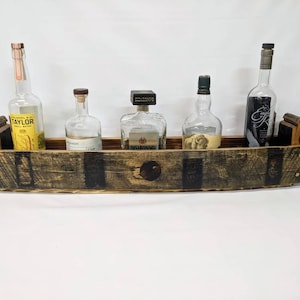 May include: A rustic wooden shelf made from reclaimed bourbon barrel staves, with five slots for displaying bottles of liquor. The shelf is stained dark brown and has a natural wood grain. The shelf is mounted on a white wall.
