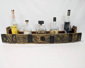 Puede incluir: Una estantería de madera rústica hecha de duelas de barril de bourbon recuperadas, con cinco ranuras para exhibir botellas de licor. La estantería está teñida de marrón oscuro y tiene una veta de madera natural. La estantería está montada en una pared blanca.