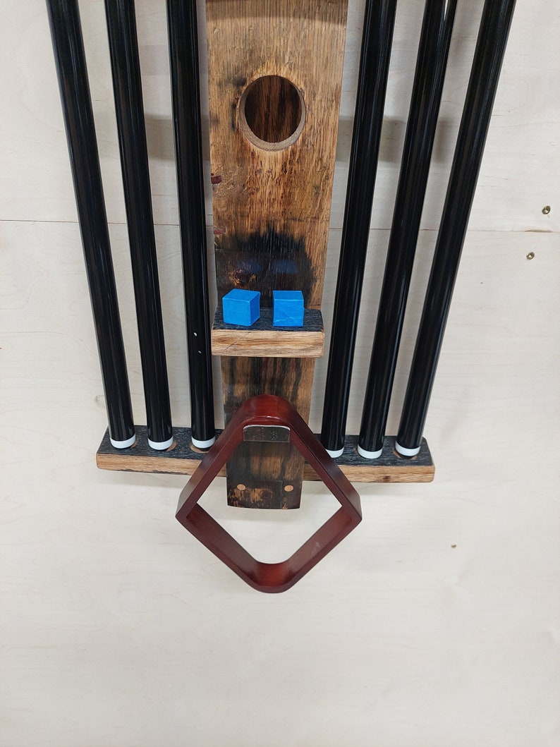May include: A wooden pool cue rack with six black pool cues and a brown leather rack. Two blue pool balls are on a shelf above the rack.