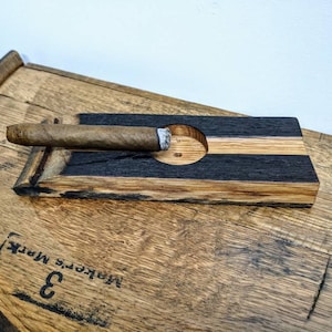 May include: A wooden cigar rest with a black and natural wood finish. The rest has a circular indentation for holding a cigar and is sitting on a wooden surface.