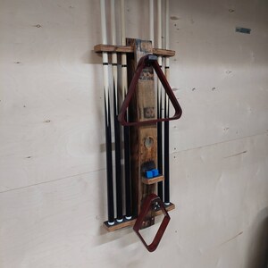 May include: A wooden wall-mounted rack for storing pool cues. The rack has a brown stain and features a black metal frame with white plastic tips. The rack also includes a red wooden triangle rack and a red wooden diamond rack.