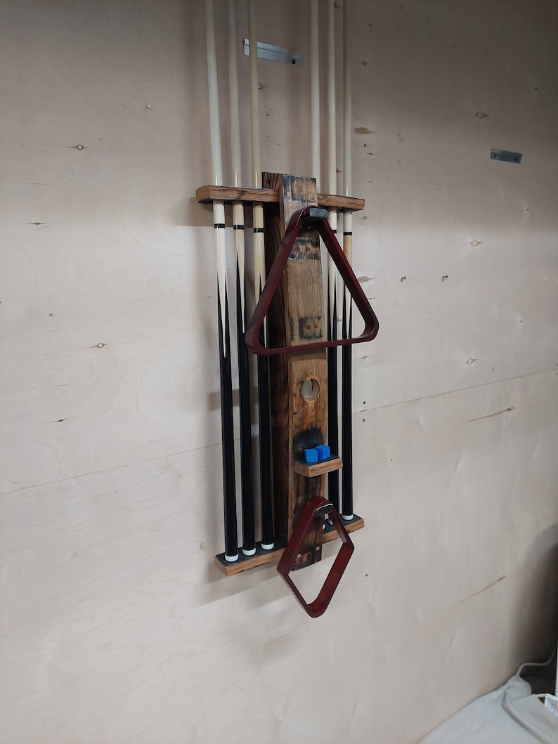 May include: A wooden wall-mounted rack for storing pool cues. The rack has a brown stain and features a black metal frame with white plastic tips. The rack also includes a red wooden triangle rack and a red wooden diamond rack.