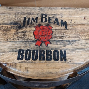 K&ouml;nnte beinhalten: Eine halbkreisf&ouml;rmige Tischplatte aus einem wiederverwendeten Fass mit dem schwarzen Schriftzug Jim Beam und Bourbon sowie einem roten kreisf&ouml;rmigen Siegel in der Mitte. Die nat&uuml;rliche Holzmaserung ist sichtbar und das St&uuml;ck hat dunkle Metallb&auml;nder.