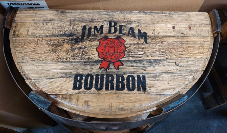 K&ouml;nnte beinhalten: Eine halbkreisf&ouml;rmige Tischplatte aus einem wiederverwendeten Fass mit dem schwarzen Schriftzug Jim Beam und Bourbon sowie einem roten kreisf&ouml;rmigen Siegel in der Mitte. Die nat&uuml;rliche Holzmaserung ist sichtbar und das St&uuml;ck hat dunkle Metallb&auml;nder.