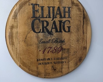 Op de afbeelding: Een rond houten bord met de tekst "Elijah Craig" in zwarte letters. Het bord bevat ook de tekst "Small Batch" en ".1789." in rode letters. De tekst "Kentucky Straight Bourbon Whiskey" is ook in zwarte letters opgenomen.