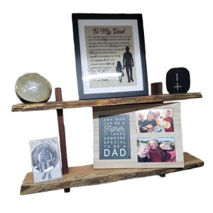 May include: A two-tiered wooden shelf with a rustic finish. The top shelf has a framed print with the text "To My Dad", a small black speaker, and a wooden object. The bottom shelf has a framed print with the text "Any man can be a father, it takes someone special to be a dad", a framed photo of a family, and a framed print with the text "I love my dad".