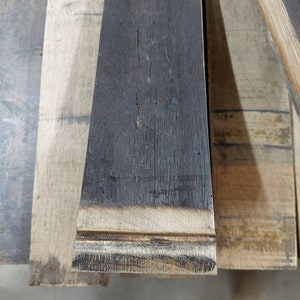 May include: Close-up of a stack of reclaimed wood planks. The top plank is dark brown and has a charred edge. The planks are all different shades of brown and have a rough, textured surface.