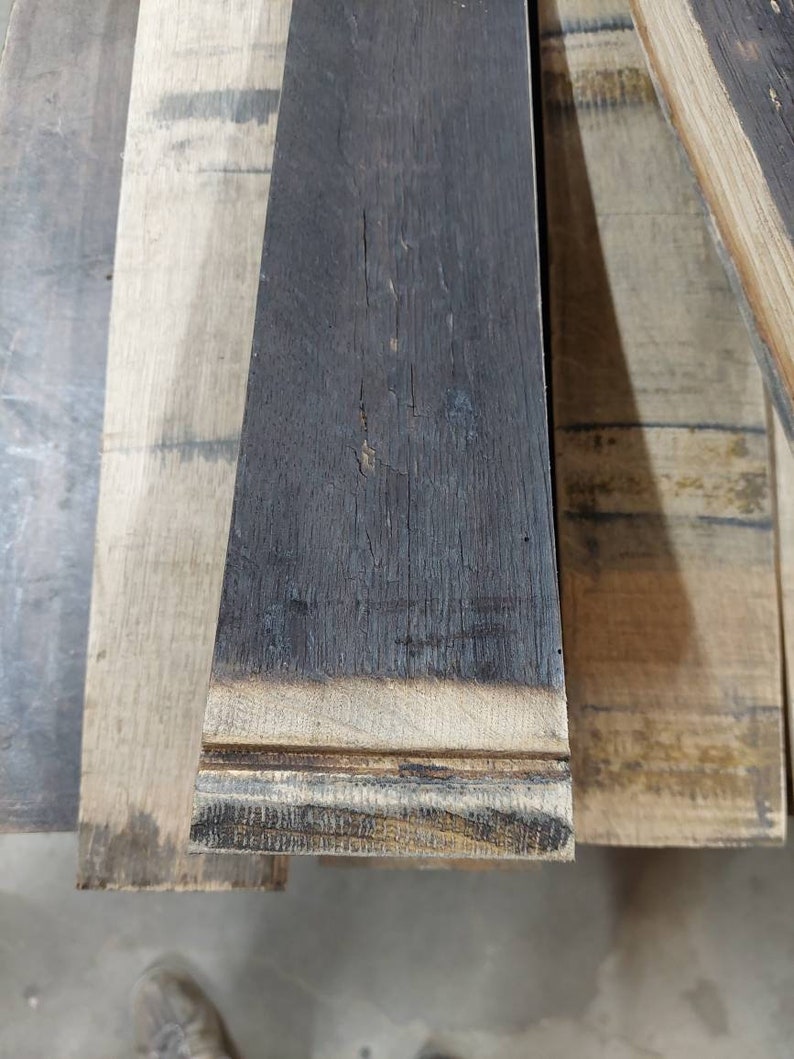 May include: Close-up of a stack of reclaimed wood planks. The top plank is dark brown and has a charred edge. The planks are all different shades of brown and have a rough, textured surface.