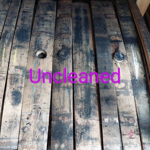 May include: Close-up of a wooden barrel stave, showing the natural wood grain and dark brown color. The stave has a circular hole and a black metal cap. The word "Uncleaned" is written in pink letters across the stave.
