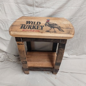 May include: A wooden end table made from a repurposed Wild Turkey Bourbon barrel. The table has a single shelf and a top with the Wild Turkey logo and a turkey illustration.