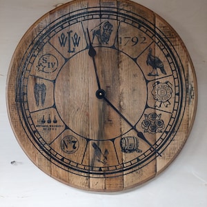 Könnte beinhalten: Ein hölzernes Zifferblatt mit schwarzen Markierungen und einem schwarzen Uhrzeiger. Das Zifferblatt zeigt verschiedene Bilder und Texte, darunter "W.L.W", "SIV", "Bourbon Whiskey", "Old No. 7 Brandy", "1792" und einen Vogel.