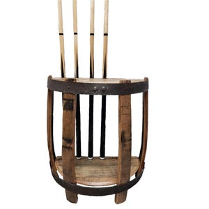 May include: A rustic wooden pool cue rack made from a repurposed barrel. The rack holds four cues, with a natural wood finish and metal bands. The cues are light-colored with black accents.