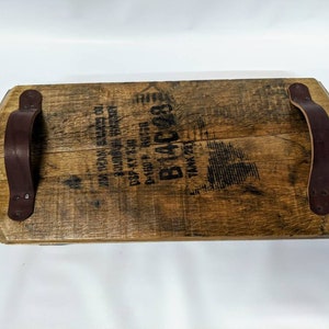 May include: A wooden serving tray made from a reclaimed whiskey barrel with two leather handles. The tray has a brown stain and the text "B&C 231" is printed on the surface.