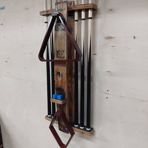 May include: A wooden wall-mounted rack for storing pool cues, with a brown stained finish. The rack has a built-in triangle rack and a small shelf for storing pool balls. The rack is mounted on a light wood wall.