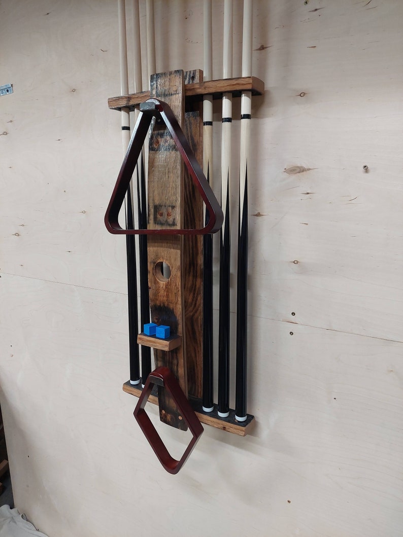 May include: A wooden wall-mounted rack for storing pool cues, with a brown stained finish. The rack has a built-in triangle rack and a small shelf for storing pool balls. The rack is mounted on a light wood wall.