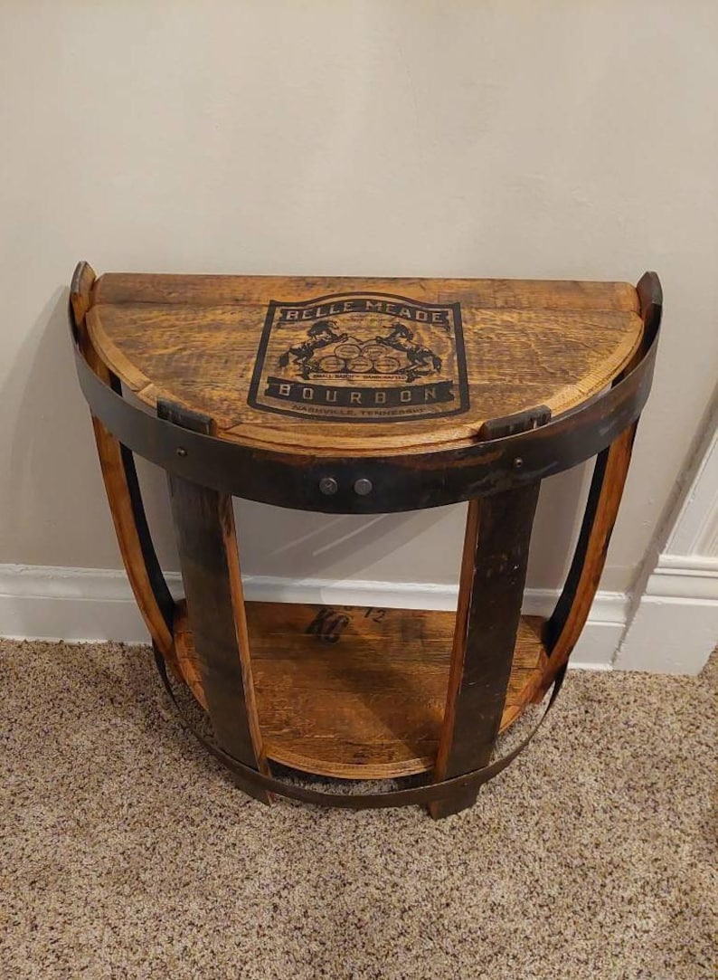 K&ouml;nnte beinhalten: Ein halbkreisf&ouml;rmiger Konsolentisch aus einem wiederverwendeten Holzfass mit dunklen Metallreifen. Die Tischplatte zeigt ein schwarzes aufgedrucktes Logo mit der Aufschrift Belle Meade Bourbon und einer Abbildung von zwei Pferden und F&auml;ssern.