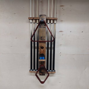 May include: A wooden wall-mounted rack for storing pool cues. The rack has a barrel design with a brown stain and a clear coat finish. It features a triangular cue rack and a diamond-shaped rack for holding a pool cue triangle. The rack is mounted on a light wood wall.