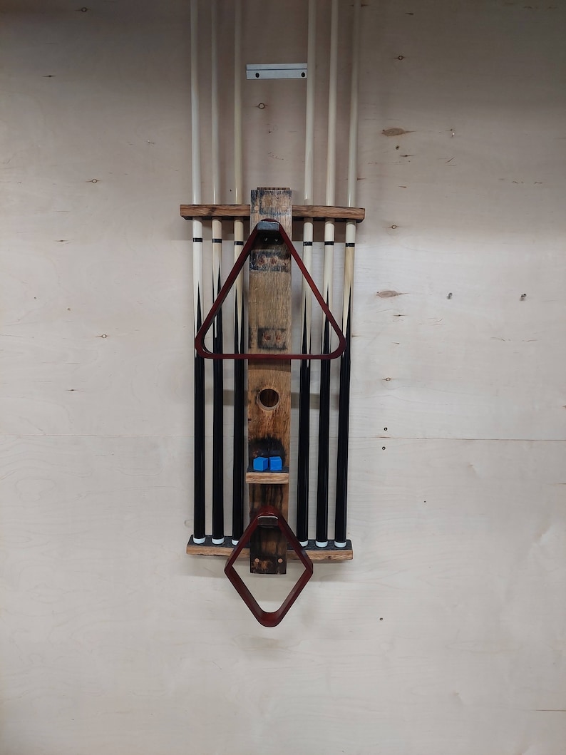May include: A wooden wall-mounted rack for storing pool cues. The rack has a barrel design with a brown stain and a clear coat finish. It features a triangular cue rack and a diamond-shaped rack for holding a pool cue triangle. The rack is mounted on a light wood wall.