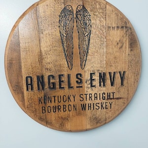 May include: A round wooden sign with the words "Angels Envy Kentucky Straight Bourbon Whiskey" and a pair of angel wings.