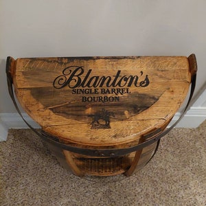K&ouml;nnte beinhalten: Ein halbkreisf&ouml;rmiges Holzregal aus einem wiederverwendeten Fass mit einer flachen Oberseite, die den schwarzen Schriftzug Blanton's SINGLE BARREL BOURBON und eine Grafik von Pferd und Jockey zeigt. Das Regal hat ein dunkles Metallband am Rand.