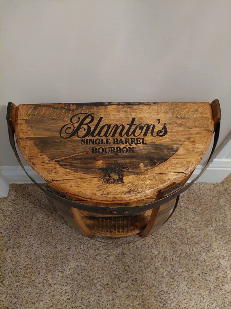 K&ouml;nnte beinhalten: Ein halbkreisf&ouml;rmiges Holzregal aus einem wiederverwendeten Fass mit einer flachen Oberseite, die den schwarzen Schriftzug Blanton's SINGLE BARREL BOURBON und eine Grafik von Pferd und Jockey zeigt. Das Regal hat ein dunkles Metallband am Rand.