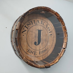 May include: A wooden barrel with a glass front, featuring the text "Nishakash Est. 1996" and the letter "J" in a circle.