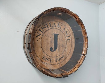 Op de afbeelding: Een houten vat met een glazen voorzijde, met de tekst "Nishakash Est. 1996" en de letter "J" in een cirkel.